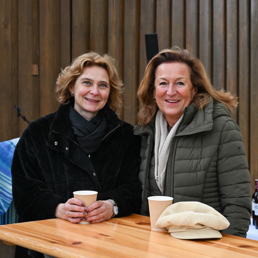 Two women sitting at a wooden table, smiling and holding cups. One wears a black coat, the other a green jacket. A beige hat and a bottle are on the table.