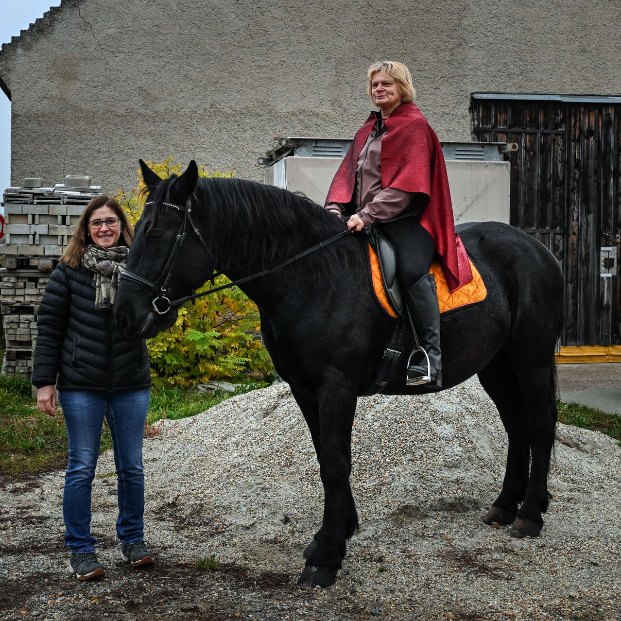 Two women stand beside a black horse. One rides the horse while the other stands next to it. They are in front of a building.