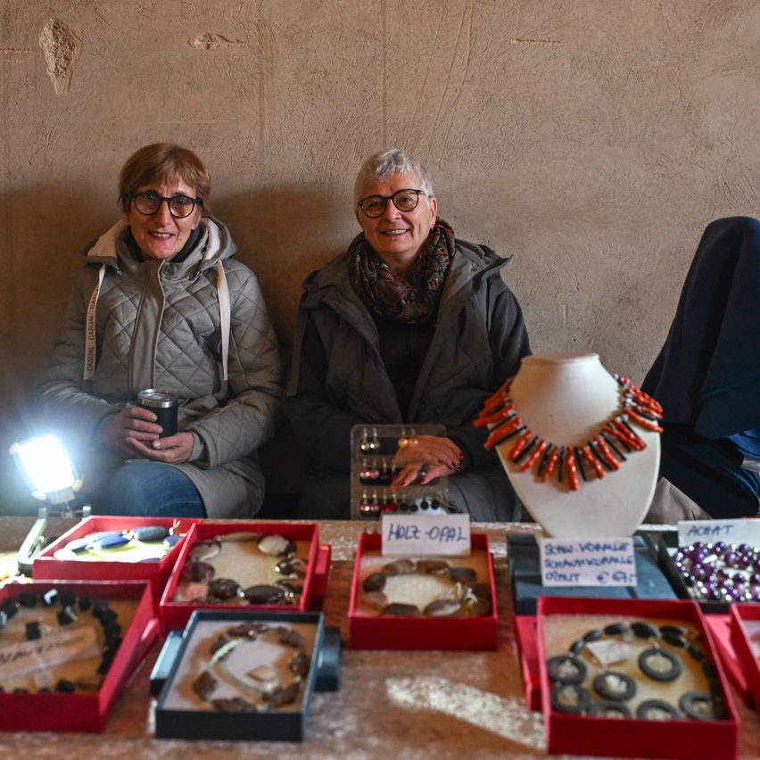 Two women are smiling and sitting next to a table with various jewelry on display. One woman holds a cup, and the table has boxes of jewelry and a sign. A lamp illuminates the scene.