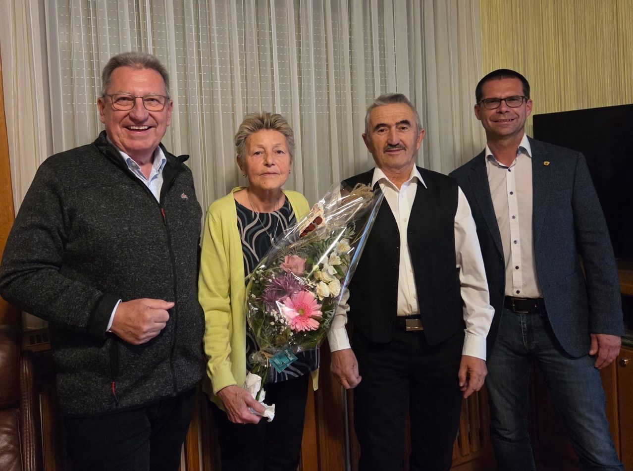Four people stand together in a room, smiling. A woman holds a bouquet of flowers. The background has white curtains.
