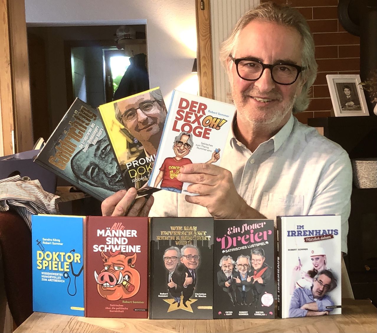 A man with glasses holds up several books in front of him. The book he holds up the most is titled Der Sex Oh! Loge. Other books include Doktor Spiele, Alle Manner Sind Schweine, and Ein Flower Dreier.