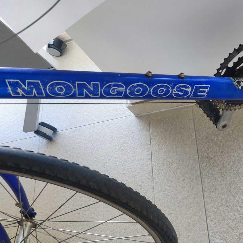 A closeup view of a blue Mongoose bicycle's frame and chain. The bicycle is on a tiled floor.