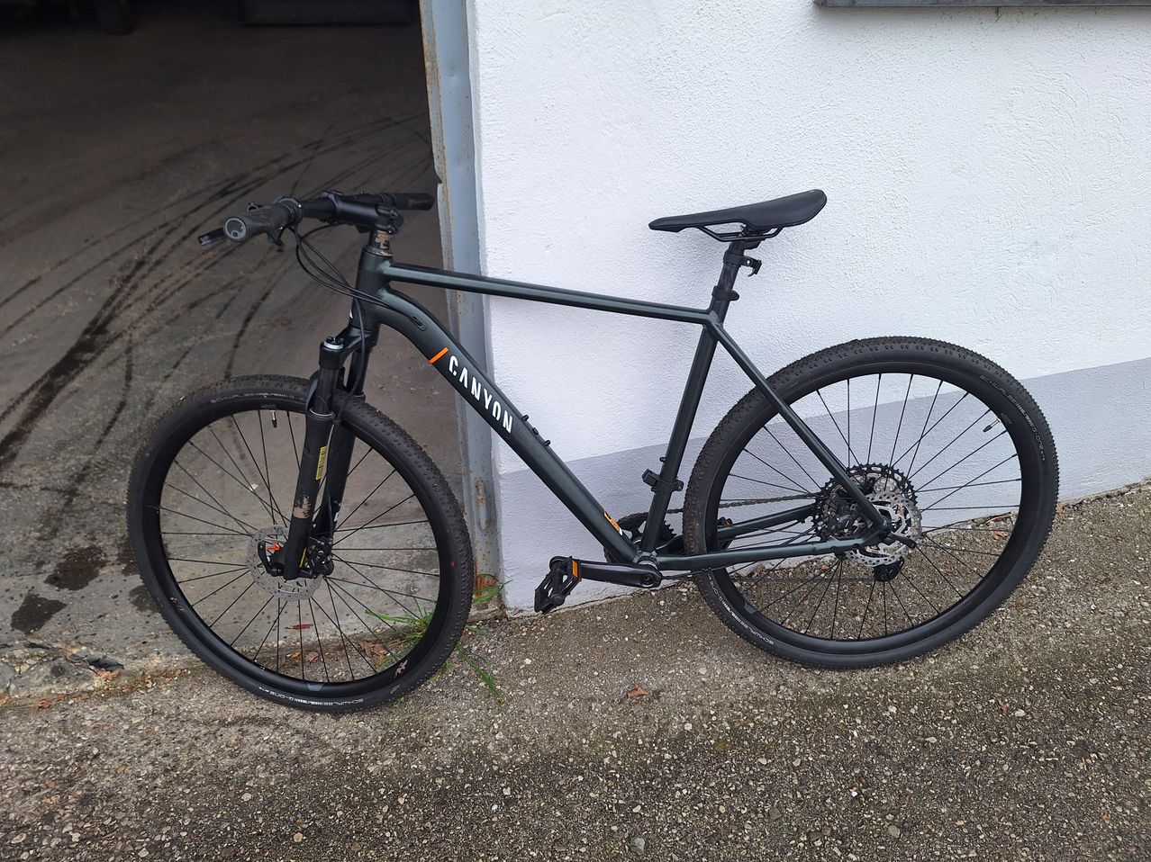A black Canyon bicycle with a black seat is parked outside a white wall. The bicycle has a disc brake, a chain, and a bottle holder.