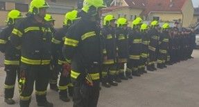 Eine Reihe von Feuerwehrleuten steht in Formation auf einer Straße. Sie tragen schwarze Uniformen mit gelben Streifen, Helmen und Stiefeln. Hinter ihnen sind mehrere Gebäude zu sehen, von denen eines ein rotes Dach hat.