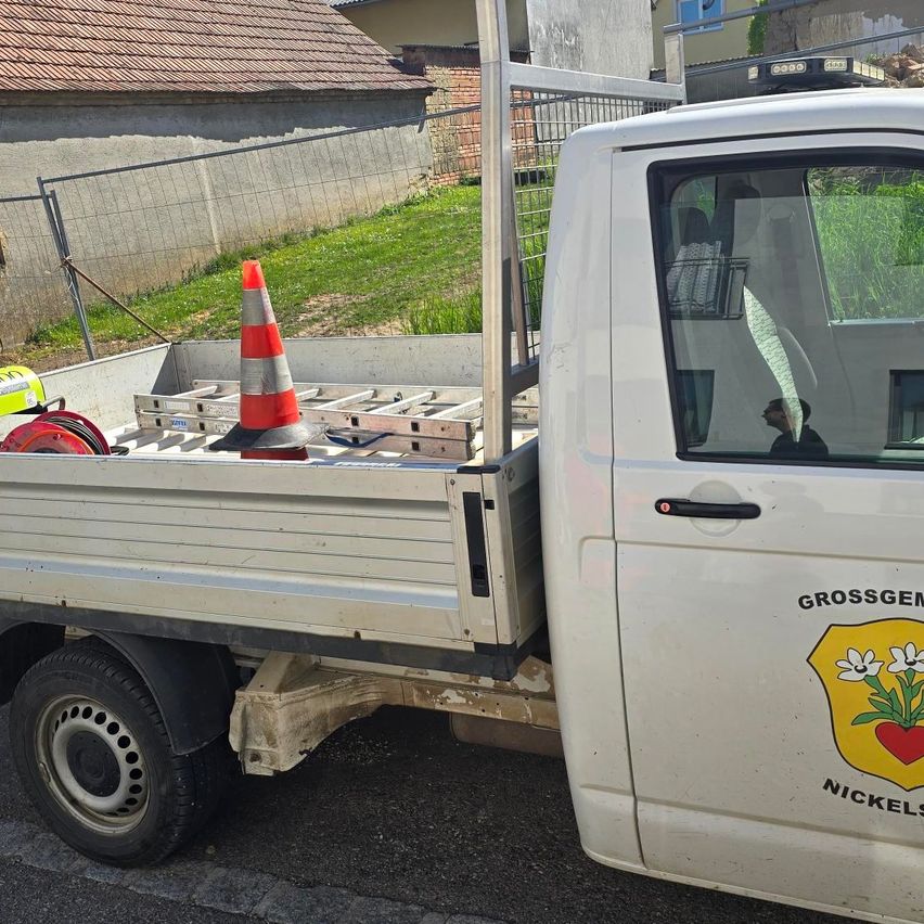 Ein weißer Pickup-Truck mit Anhänger, geparkt in der Nähe einer Mauer. Im Inneren sitzt eine Person auf dem Fahrersitz. Der Truck hat ein Logo und einen Warnkegel im Heck.