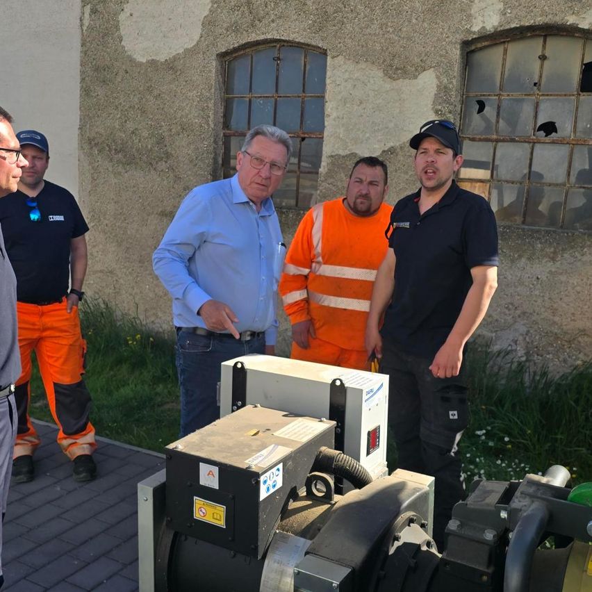 Fünf Männer stehen vor einem alten Gebäude mit zerbrochenen Fenstern. Ein Mann zeigt auf eine Maschine, während die anderen zusehen.