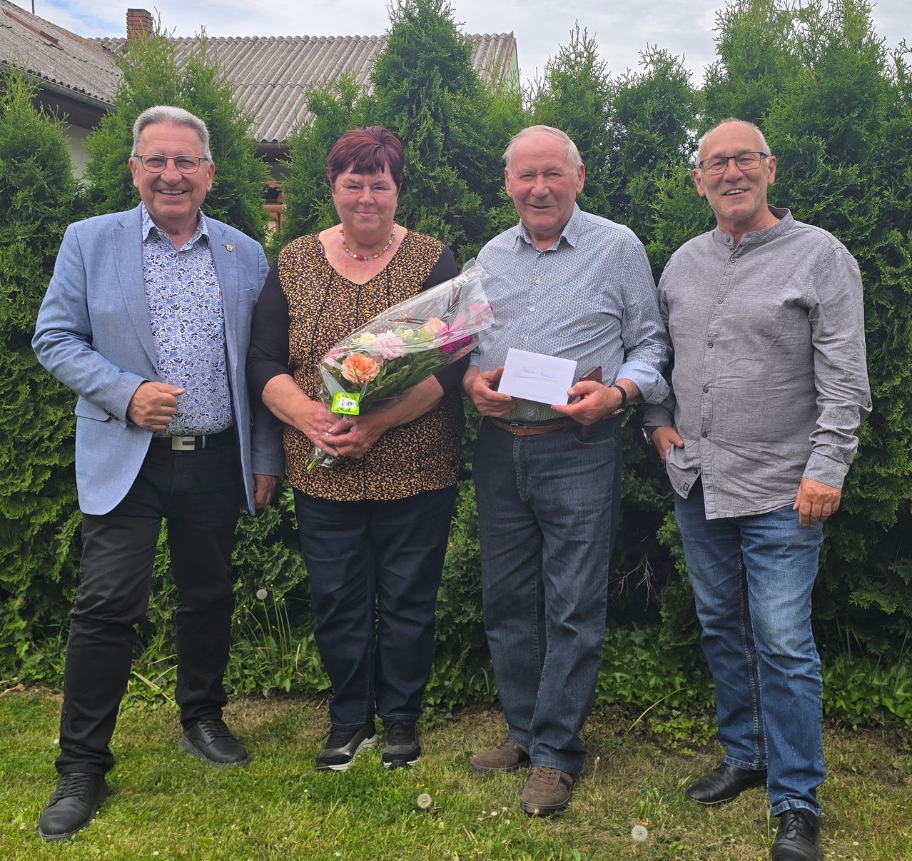 Four people stand in a garden, smiling and posing for a photo. A woman holds a bouquet of flowers, and a man holds a paper. Behind them are bushes.