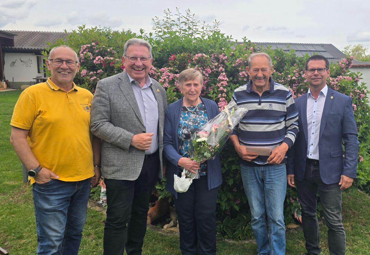 Fünf Menschen stehen lächelnd vor einem Busch. Die Frau in der Mitte hält einen Blumenstrauß. Dahinter ist ein Garten mit verschiedenen Pflanzen und Blumen zu sehen.