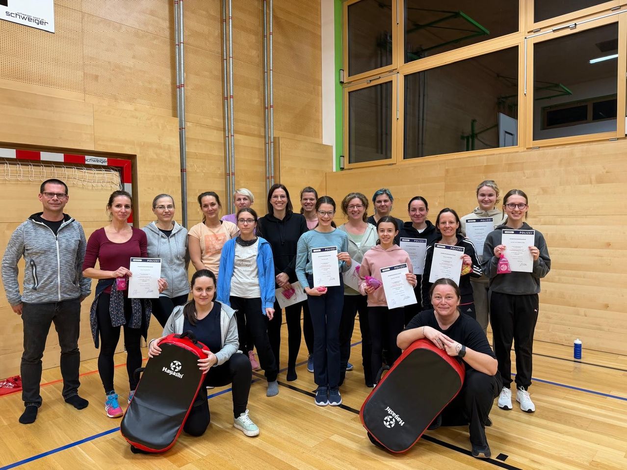A group of women pose for a photo in a gym, holding certificates, with two individuals kneeling on the floor with sports bags.