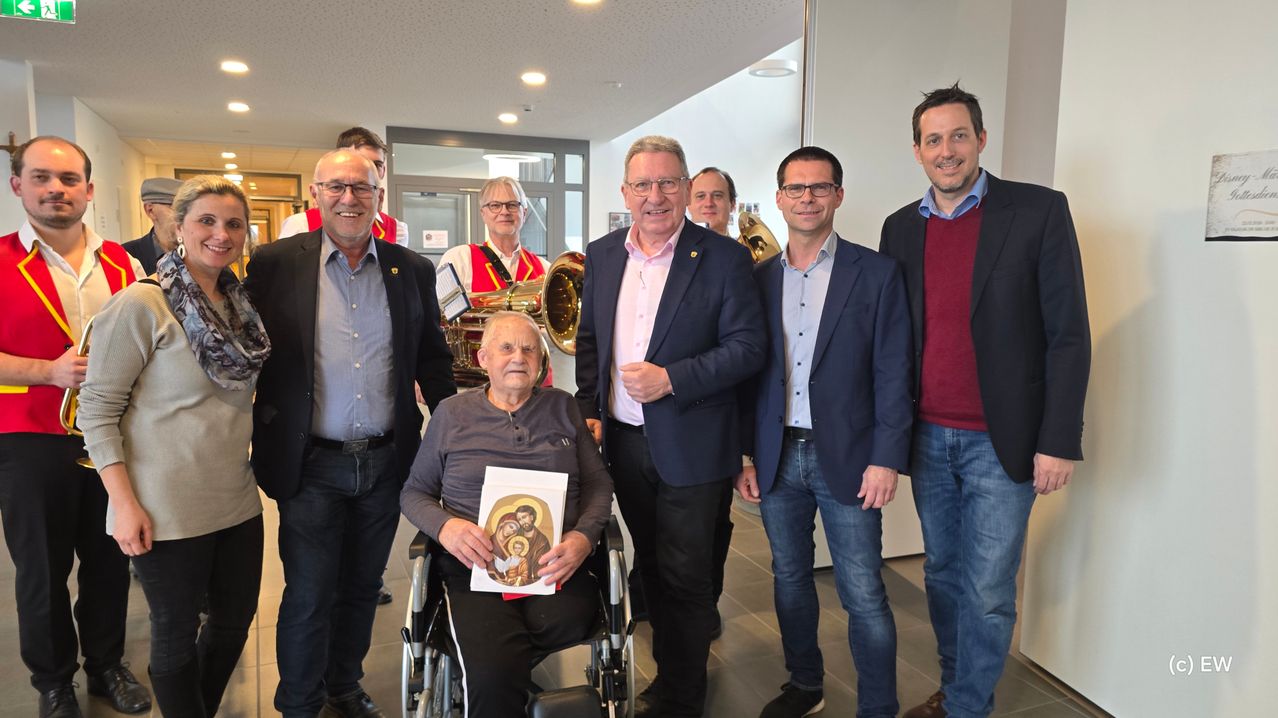 Eine Gruppe von Männern steht in einem Flur mit einer Person im Rollstuhl, die ein religiöses Bild hält, Brille trägt. Dahinter tragen zwei Personen Musikinstrumente.