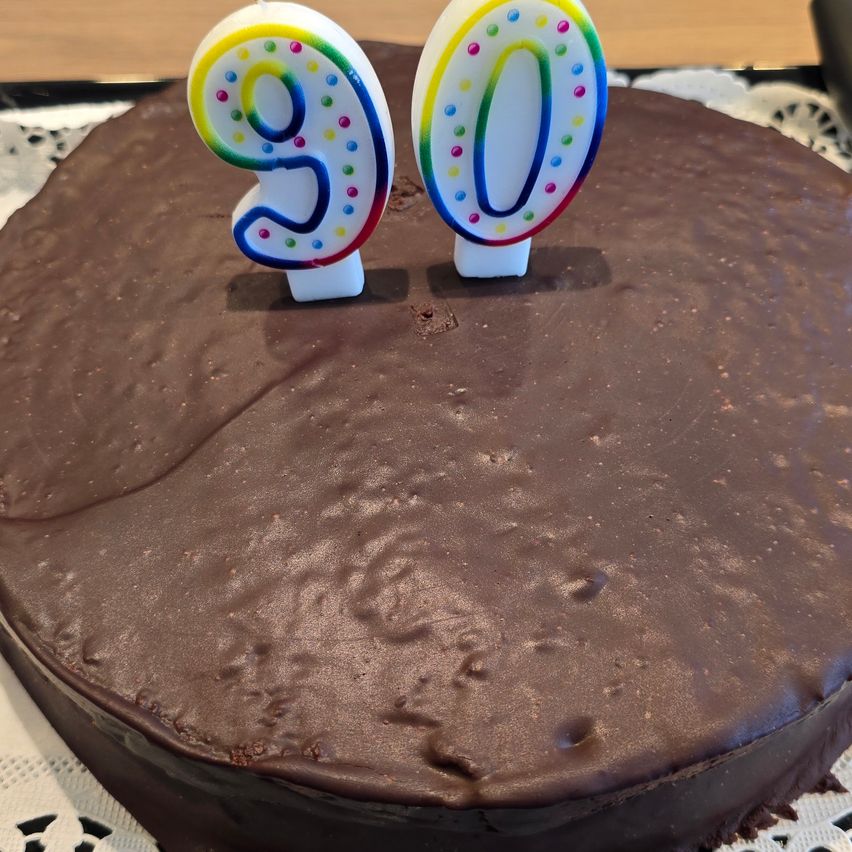 Ein Schokoladenkuchen mit zwei Kerzen, eine in Form einer 9 und die andere in Form einer 0, steht auf einem weißen Doily. Der Kuchen hat eine glänzende Oberfläche.