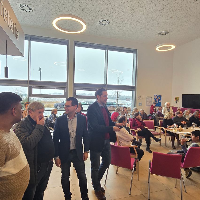 Eine Gruppe von Menschen in formeller Kleidung versammelt sich in einem Speisebereich mit großen Fenstern. Zwei Männer gehen auf einen Tisch zu, während andere an Tischen sitzen, essen und plaudern.