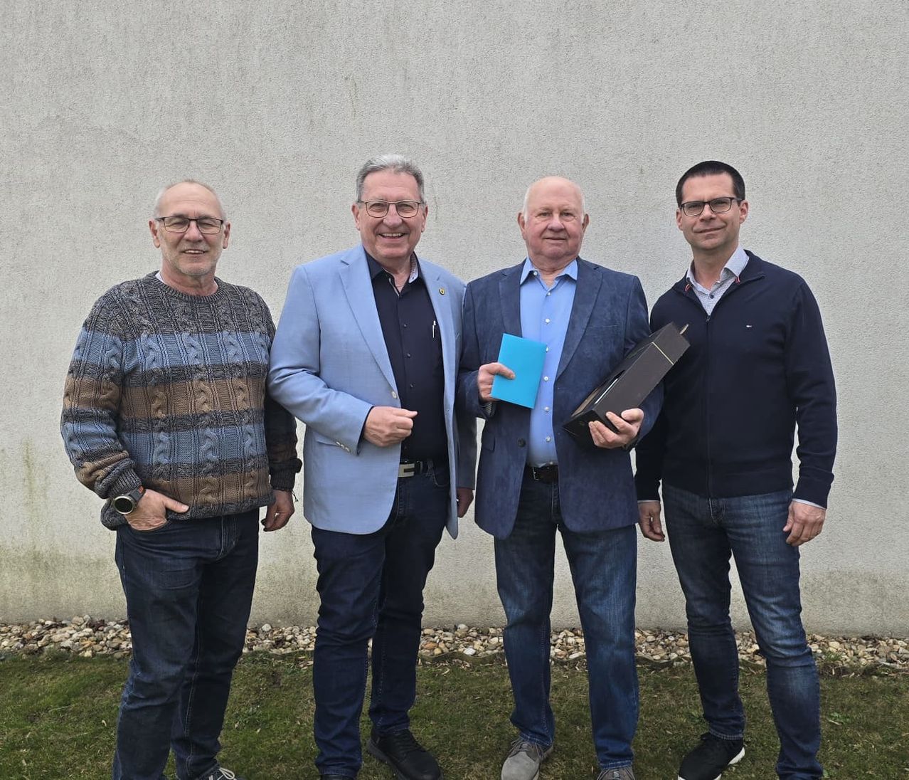 Vier Männer stehen vor einem Gebäude und lächeln für ein Foto. Ein Mann hält eine blaue Mappe, ein anderer einen Kasten und der Mann in der Mitte trägt einen Ausweis.