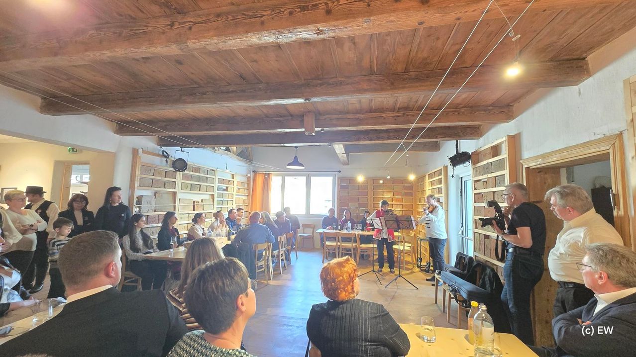 Eine Gruppe von Menschen ist in einem Raum mit Holzdecken und -wänden versammelt. Ein Violinist und ein Gitarrist treten auf der Bühne auf. Das Publikum sitzt an Tischen und Stühlen. Die Bühne ist mit Musikinstrumenten und einem Mikrofon ausgestattet. An der Wand befinden sich auch einige Gegenstände.