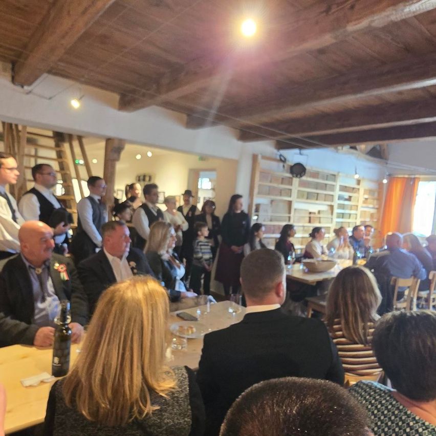 Eine große Gruppe von Menschen versammelt sich in einem Raum mit Holzbalken, Tischen und Stühlen. Einige stehen, andere sitzen. Eine Flasche, Teller und Gläser stehen auf dem Tisch. Einige tragen Hüte, Brillen und Mäntel. Eine Leiter lehnt an der Wand.