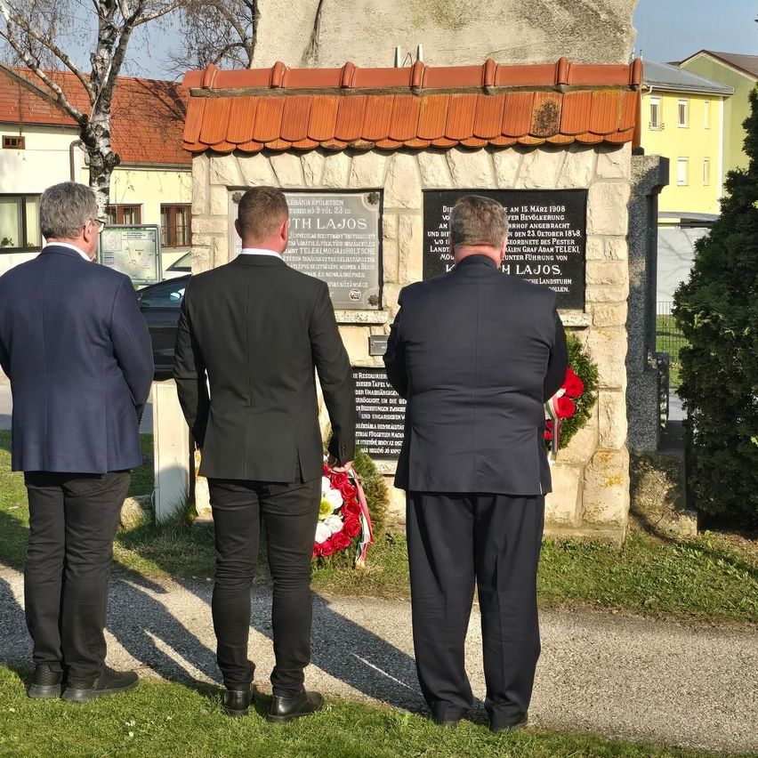 Drei Männer in Anzügen stehen vor einem Gedenkstein mit einem Blumenkranz. Der Mann auf der rechten Seite schaut auf den Stein, während die anderen den Kranz betrachten.