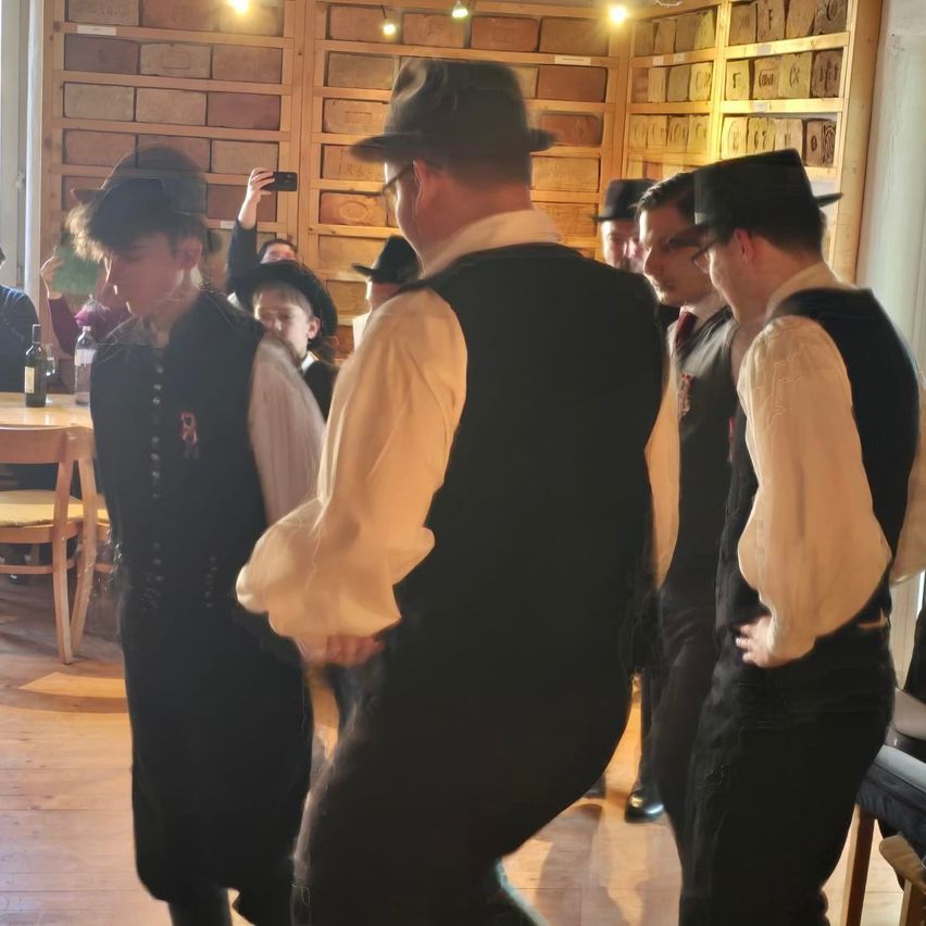 Eine Gruppe von Menschen in Vintage-Kleidung, möglicherweise bei einer historischen Nachstellung, steht in einem Raum. Einige tragen Hüte und Brillen. Ein Tisch und Stühle sind in der Nähe.