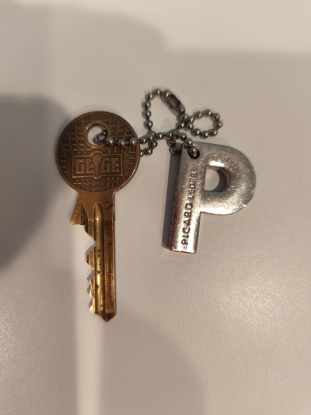 A brass key with a lock design and a silver pendant with the letter P and the word Ricard on it.