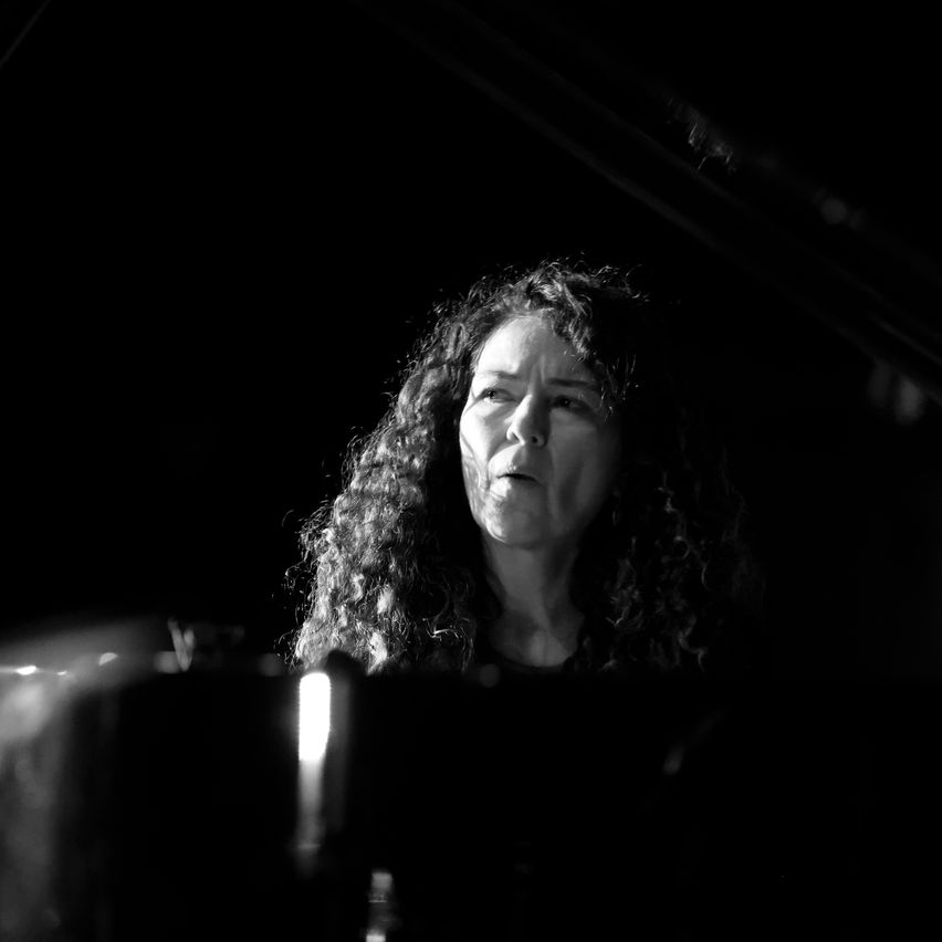 Ein Schwarz-Weiß-Foto einer Frau mit lockigem Haar, die Klavier spielt, ihr Gesicht von einem Scheinwerfer beleuchtet.
