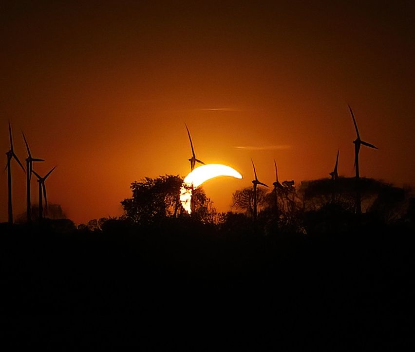 A partial solar eclipse at sunset with wind turbines in silhouette against the orange sky.