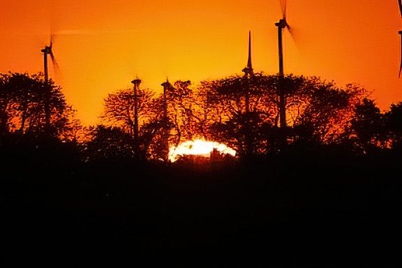 A silhouette of wind turbines against a fiery orange sunset sky with a fire burning in the foreground.