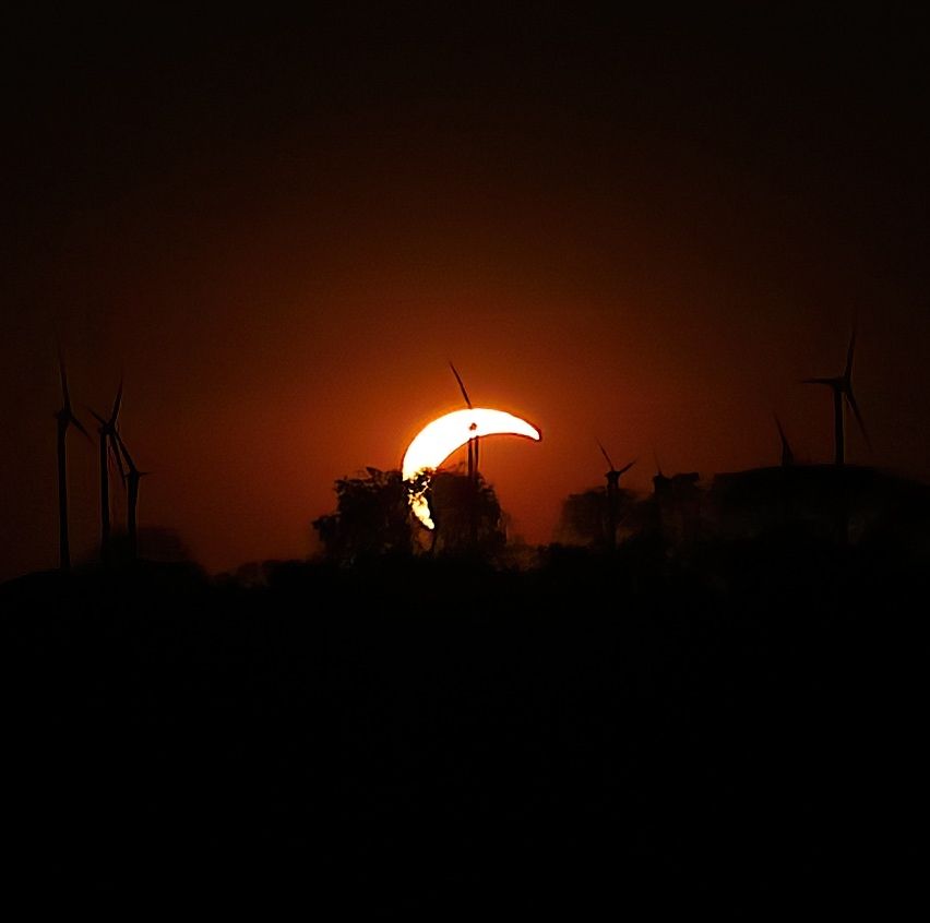 A partial solar eclipse with a crescent moon visible against a dark sky, silhouetted by wind turbines.