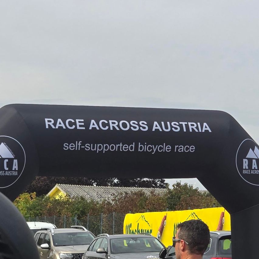 Ein Mann steht unter einem schwarzen Bogen mit der Aufschrift 'RACE ACROSS AUSTRIA' und einem Logo für ein selbst unterstütztes Fahrradrennen, mit Autos und einem gelben Banner im Hintergrund.
