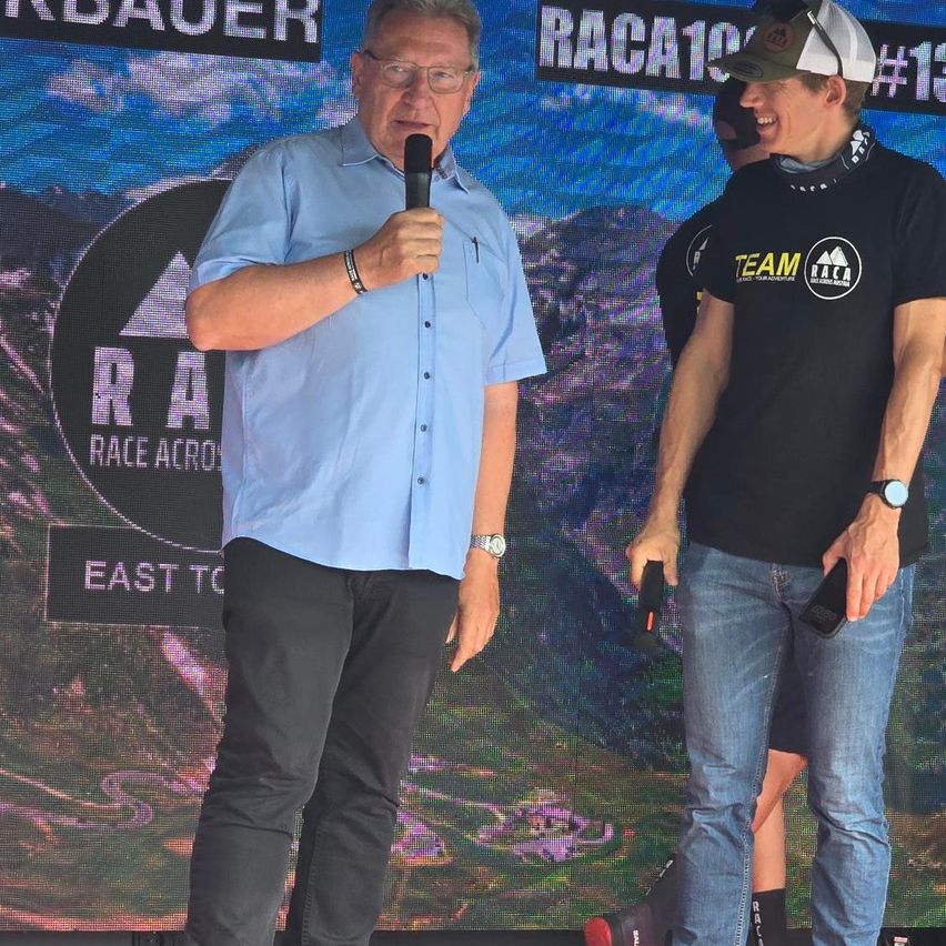 Zwei Männer stehen auf einer Bühne mit einer großen Leinwand im Hintergrund. Der Mann auf der linken Seite hält ein Mikrofon und trägt ein hellblaues Hemd. Der Mann auf der rechten Seite trägt ein schwarzes T-Shirt mit weißem Text und eine Kappe.