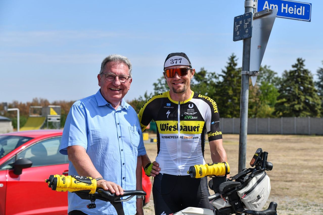 Zwei Männer stehen neben einem Fahrrad, einer trägt ein Radtrikot mit der Nummer 377, der andere ein blaues Hemd. Sie lächeln und scheinen für ein Foto zu posieren. Dahinter steht ein Pfosten mit einem Schild und ein rotes Auto.