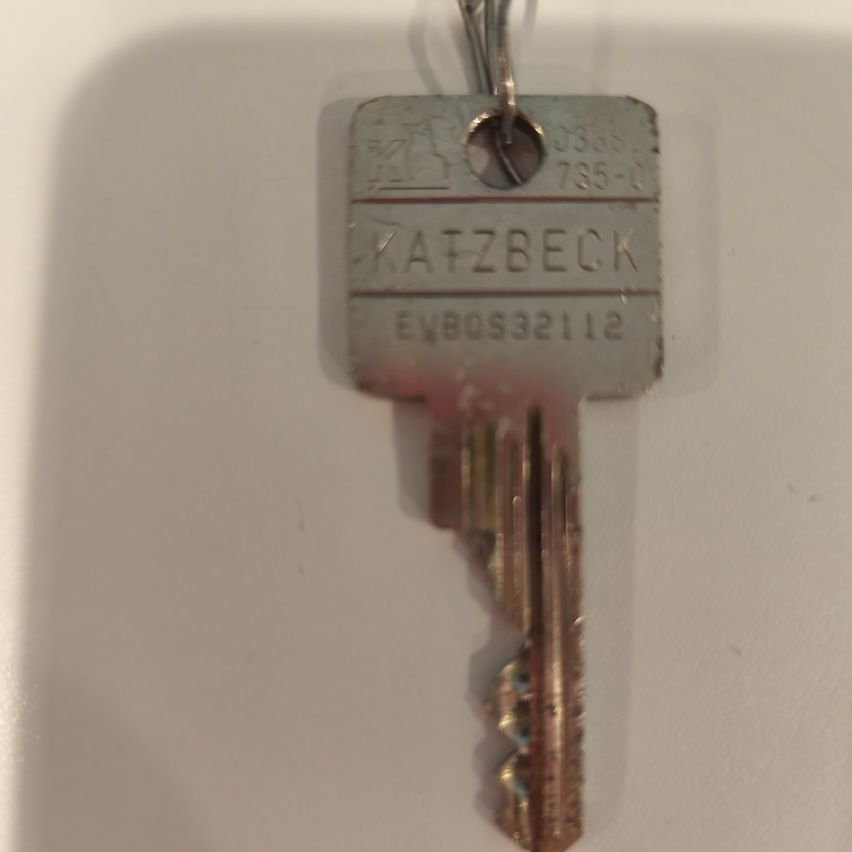 Ein silberner Schlüssel mit der Aufschrift 'KATZBECK' und der Nummer '735-0' hängt an einer weißen Oberfläche.