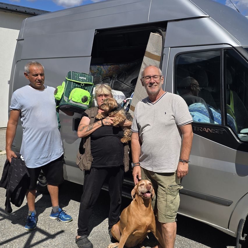 Drei Personen und zwei Hunde stehen vor einem Van, wobei eine Person einen kleinen Hund und eine andere eine Hundetasche hält.