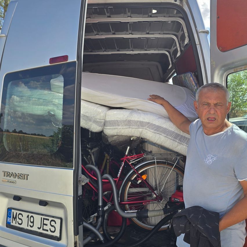 Ein Mann steht neben einem Van mit offener Heckklappe, die eine Matratze und ein Fahrrad im Inneren zeigt. Der Van hat ein Kennzeichen mit der Aufschrift MS 19 JES.