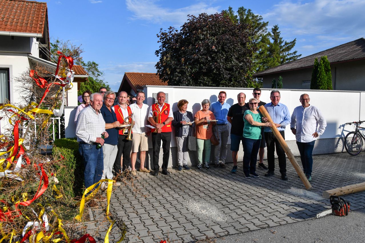 Eine Gruppe von Menschen steht vor einer weißen Wand mit einem Haus und Bäumen im Hintergrund. Sie halten Musikinstrumente und tragen Freizeitkleidung.