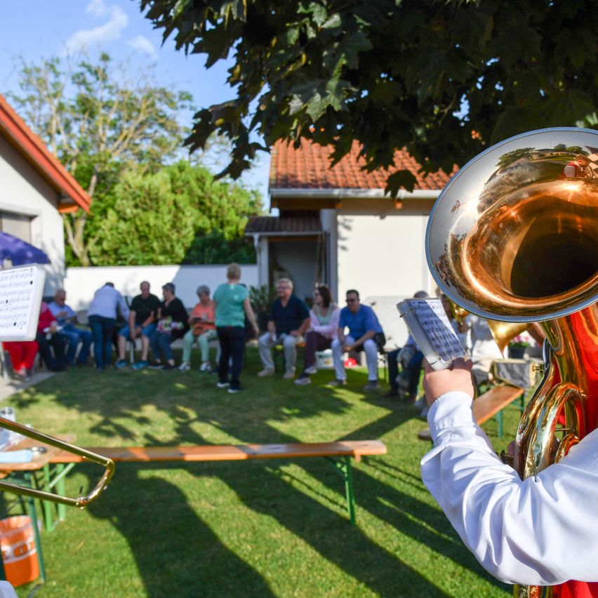 Eine Person spielt ein Blasinstrument im Hinterhof eines Hauses, vor einer sitzenden Zuhörerschaft. Die Person hält ein Notenblatt.
