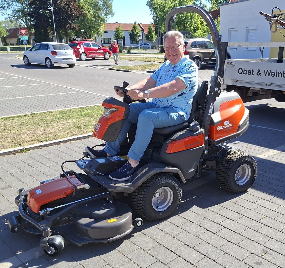 A man sits on a Husqvarna mower, smiling, with a white truck parked behind him. Cars are parked in a parking lot.