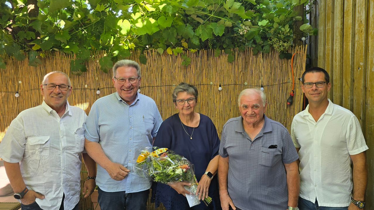 Three people standing in front of a bamboo wall, one holding a bouquet of flowers. They are smiling and seem to be posing for a photo.