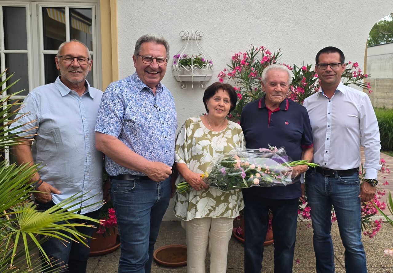 Five adults stand outside, smiling for a photo. A woman holds a bouquet of flowers. Behind them are potted plants.