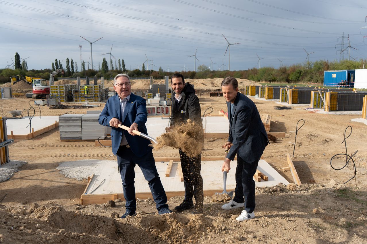 Drei Männer in Anzügen sind an einer Baustelle, jeder hält eine Schaufel und wirft Erde. Die Baustelle ist von Holzkonstruktionen und Maschinen umgeben. Im Hintergrund befinden sich mehrere Windturbinen und Bäume.