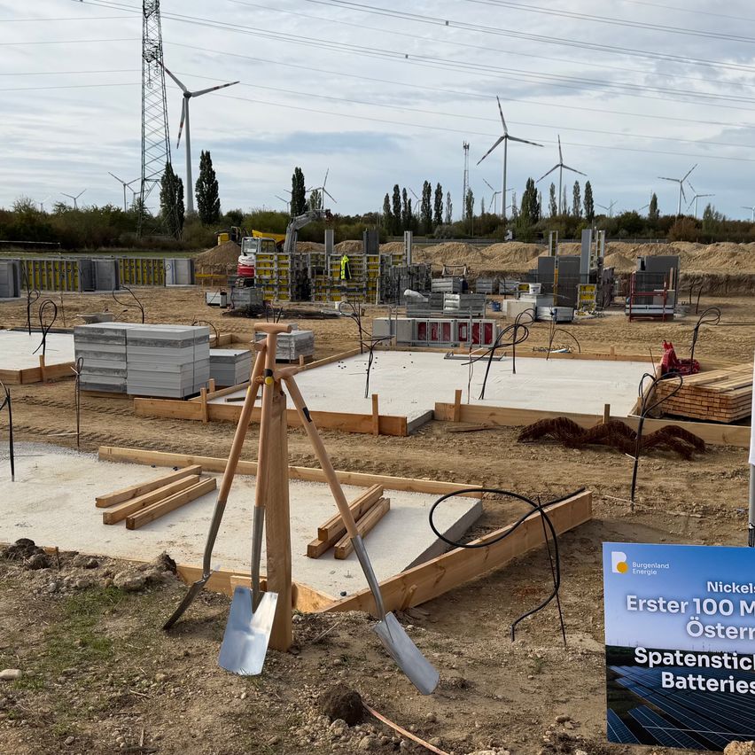 Eine Baustelle mit Schaufeln und einem Schild für Nickel 100 MW Österreich Spatenstich Batteries. Der Standort ist von Erde, Holzbrettern und verschiedenen Baumaterialien umgeben. Im Hintergrund befinden sich mehrere Windräder und Bäume.