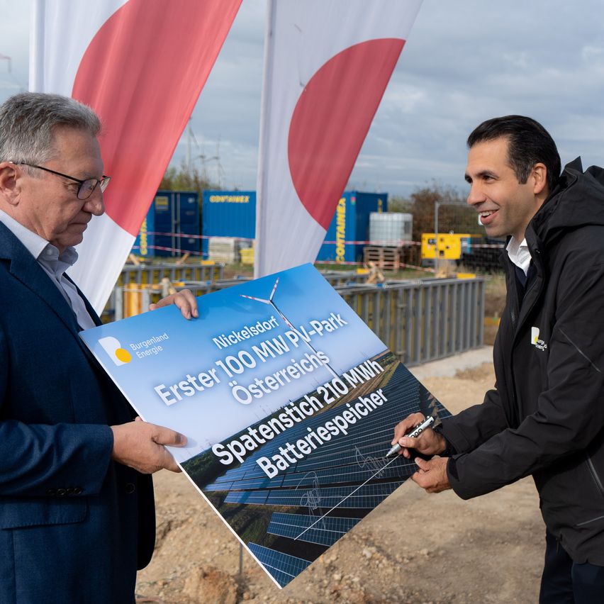 Zwei Männer in formeller Kleidung halten ein großes Schild an einer Baustelle. Das Schild steht 'Erster 100 MW PV-Park Osterreich 210 MWh' mit Solarpanelen im Hintergrund.