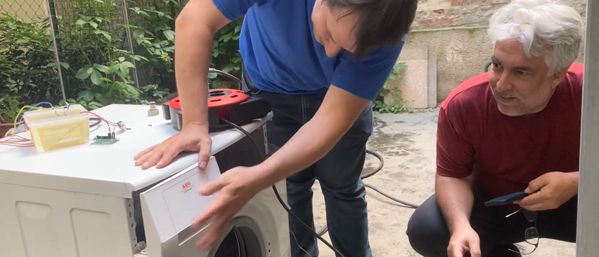 Ein Mann in einem blauen T-Shirt repariert eine weiße Waschmaschine, während eine andere Person in einem roten T-Shirt neben ihm kniet.