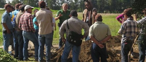 Eine Gruppe von Menschen in Freizeitkleidung steht auf einem Feld, einige graben, während andere beobachten. Eine Person mit einer Tasche steht in der Nähe.