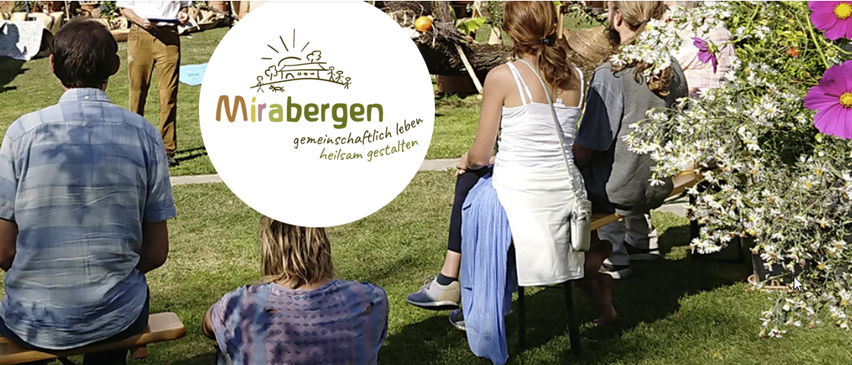 Eine Gruppe von Menschen sitzt auf Hockern in einem Gartenbereich. Eine Person trägt ein weißes Kleid, und die anderen tragen Freizeitkleidung. Ein Mann in braunen Hosen steht hinter ihnen. Es gibt Blumen und Pflanzen im Bereich.
