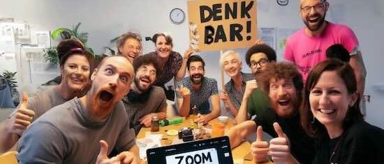 Eine Gruppe von Menschen sitzt um einen Tisch herum, lächelt und gibt Daumen hoch. Ein Laptop mit der Zoom-App ist auf dem Tisch geöffnet.