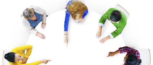 Eine Gruppe von Menschen sitzt um einen runden Tisch herum, jeder mit unterschiedlicher Kleidung und in verschiedenen Gesten, was auf eine lebhafte Diskussion hindeutet.