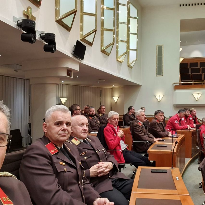 Militäroffiziere in formellen Uniformen sitzen in einem Konferenzraum, einige klatschen. Eine Person in Rot steht in der Nähe der Vorderseite. Mikrofone und Schreibtische sind vorhanden.