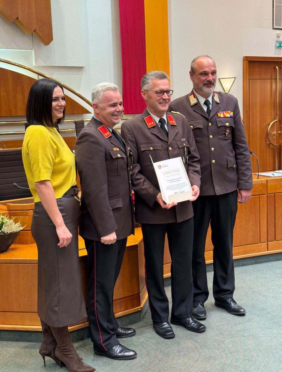 Drei Männer in Militäruniformen und eine Frau in einem gelben Oberteil stehen vor einem Podium, lächeln und halten eine Urkunde. Hinter ihnen hängt eine rote Fahne an der Wand.