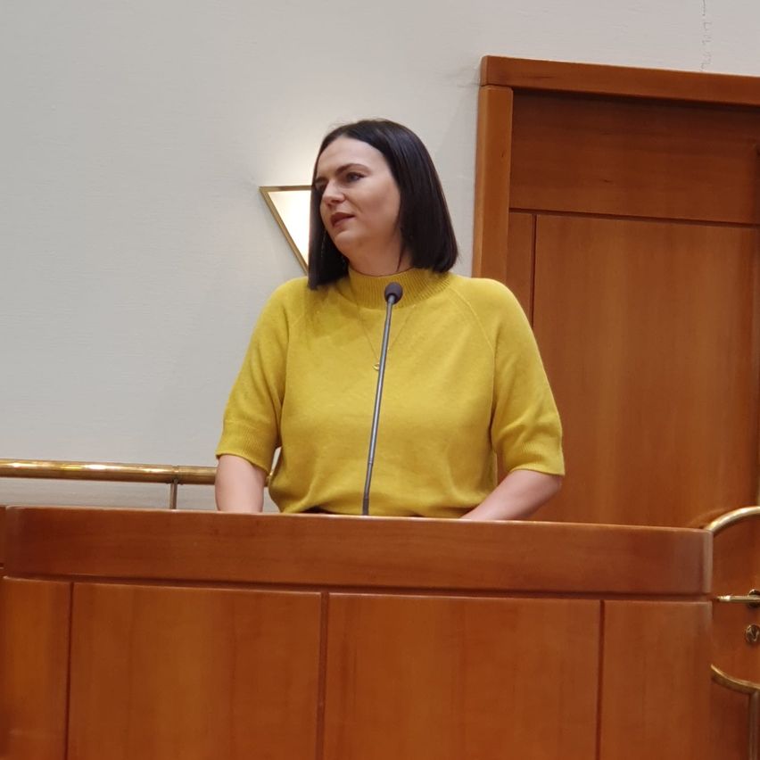 Eine Frau in einem gelben Pullover steht an einem Rednerpult mit einem Mikrofon in einem Raum mit Holzwänden.