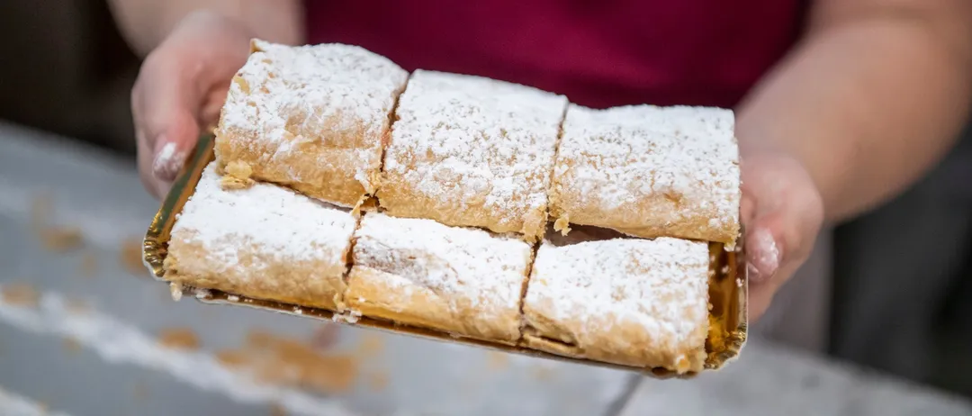 Eine Person hält eine Schale mit sechs Stücken Gebäck, bestreut mit Puderzucker, wahrscheinlich in einer Bäckerei.