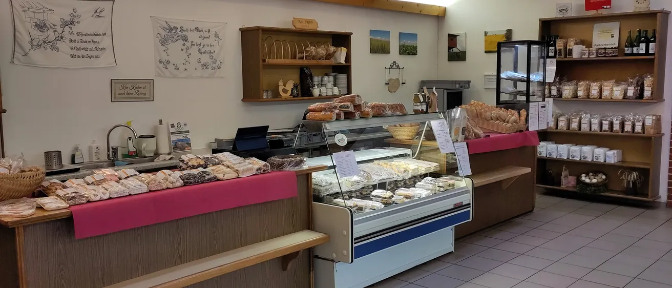 Eine Bäckerei mit Holzregalen, gefüllt mit Brot und Gebäck. Ein Ausstellungsfall mit verschiedenen Desserts steht auf dem Tresen, und ein weiteres Regal hält mehr Brot. Verschiedene Gegenstände hängen an der Wand.